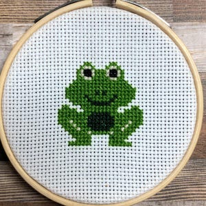 May include: A cross-stitch embroidery hoop featuring a green frog design. The frog is centered on white fabric, with a wooden hoop and metal tightening mechanism. The background is a wood-grain pattern.