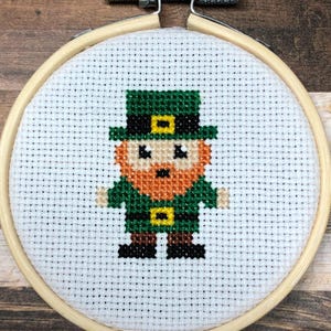Könnte beinhalten: Kreuzstich eines Kobolds in einem hölzernen Stickrahmen. Der Kobold hat einen grünen Hut, eine Jacke und Schuhe, einen roten Bart und schwarze Augen. Der Hintergrund ist weiß mit einem Rastermuster.