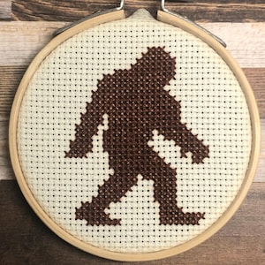 Könnte beinhalten: Ein Kreuzstich-Stickrahmen mit einer braunen Silhouette von Bigfoot. Das Design ist auf cremefarbenem Stoff gestickt und in einem hölzernen Stickrahmen eingefasst. Der Hintergrund ist eine Holzoberfläche.
