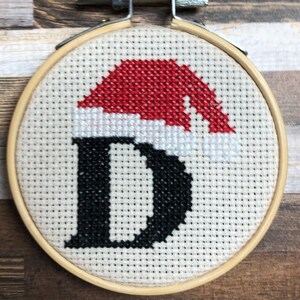 May include: A cross-stitch embroidery hoop featuring a black letter "D" under a red Santa hat. The design is on a cream-colored fabric, set in a wooden hoop. The background is a wooden surface.