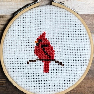 Op de afbeelding: Kruissteekborduurwerk van een rode kardinaalvogel die op een bruine tak zit. De vogel is gecentreerd op een witte stof in een houten borduurring. De ring is vastgezet met een metalen sluiting en schroef.