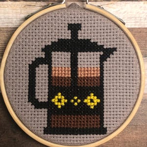 Könnte beinhalten: Kreuzstich-Stickerei einer French-Press-Kaffeemaschine in einem hölzernen Stickrahmen. Die Kaffeepresse ist schwarz mit braunen und gelben Details, vor einem grauen Hintergrund. Der Rahmen ist aus Holz.