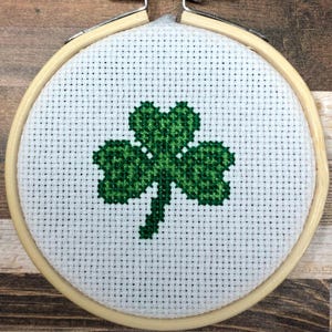 Puede incluir: Un bastidor de bordado de punto de cruz con un diseño de trébol verde sobre tela blanca. El trébol está centrado dentro del aro redondo, que es de madera clara. El aro se asegura con un tornillo y una tuerca metálicos.