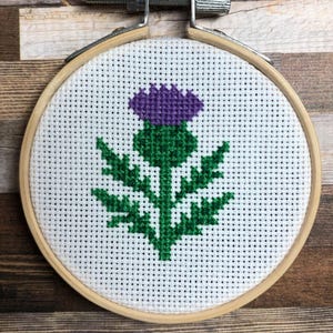 Könnte beinhalten: Kreuzstich-Stickerei einer lila Distelblume mit grünen Blättern und Stiel, vor weißem Hintergrund. Die Stickerei ist in einem Holzreifen mit einem Metallspannmechanismus gerahmt. Der Hintergrund ist eine Holzoberfläche.