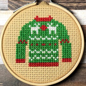 Könnte beinhalten: Kreuzstich-Stickerei eines grünen Weihnachtspullover-Designs mit weißen Rentieren und roten Akzenten, in einem hölzernen Stickrahmen. Die Handarbeit ist auf einem beigefarbenen Stoffhintergrund mit einem Holzgrund befestigt.