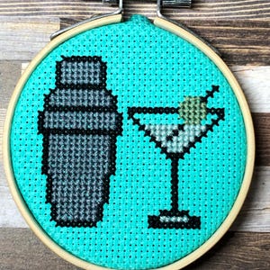 May include: Cross-stitch embroidery in a wooden hoop featuring a cocktail shaker and a martini glass with an olive. The background is a bright teal color. The embroidery is secured with a metal and wood hoop.