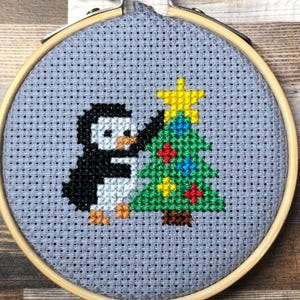 Könnte beinhalten: Kreuzstich-Stickerei in einem Holzrahmen mit einem Pinguin, der einen Weihnachtsbaum schmückt. Der Pinguin ist schwarz und weiß mit orangefarbenen Füßen. Der Baum ist grün mit bunten Ornamenten und einem gelben Stern. Der Hintergrund ist grau.