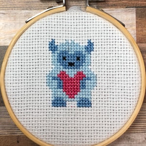 Könnte beinhalten: Kreuzstich-Stickerei in einem Holzrahmen mit einem blauen Bären, der ein rotes Herz hält. Der Bär ist auf weißem Stoff gestickt. Der Rahmen wird mit einer Metallschraube und -mutter befestigt.