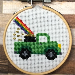 Könnte beinhalten: Kreuzstich-Stickerei in einem Holzrahmen zeigt einen grünen Lastwagen mit einem Regenbogen und Goldmünzen. Der Regenbogen erstreckt sich von der Ladefläche des Lastwagens. Die Stickerei befindet sich auf weißem Stoff, der Rahmen wird mit einer Metallschraube befestigt.