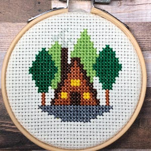 Könnte beinhalten: Fertige Kreuzstich-Stickerei in einem Holzrahmen. Das Design zeigt ein braunes Haus mit gelben Fenstern, grüne Bäume und eine graue Basis. Der Hintergrund ist weiß mit einem Rastermuster.