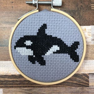 Könnte beinhalten: Ein Kreuzstich-Stickrahmen mit einem schwarz-weißen Orca-Design auf grauem Hintergrund. Der Orca befindet sich in der Mitte des runden Reifens, der mit einer Metallschraube und einem Holzrahmen befestigt ist. Der Hintergrund ist ein hellgrauer Stoff.
