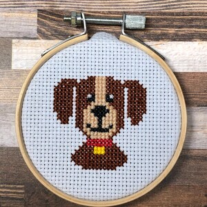 Könnte beinhalten: Kreuzstich-Stickerei eines Hundes in einem Holzreifen. Der Hund hat braunes Fell, eine hellbraune Schnauze und ein rotes Halsband mit gelbem Akzent. Der Hintergrund ist weiß mit gleichmäßig verteilten Löchern für den Kreuzstich.