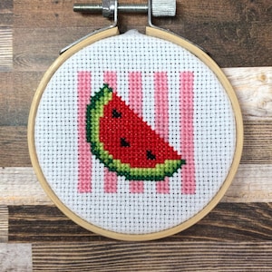 Könnte beinhalten: Ein Kreuzstich-Stickrahmen mit einem Wassermelonen-Scheiben-Design. Die Wassermelone ist rot mit schwarzen Kernen und einer grünen Schale, vor einem Hintergrund aus rosa Streifen. Der Rahmen ist aus Holz und hat einen Metallspannmechanismus.