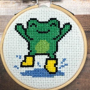 Könnte beinhalten: Kreuzstich-Stickerei einer grünen Frosch mit gelben Stiefeln, der in eine blaue Pfütze springt. Der Frosch ist auf weißem Stoff zentriert, der von einem hölzernen Stickrahmen umrahmt wird. Der Frosch hat einen fröhlichen Ausdruck.