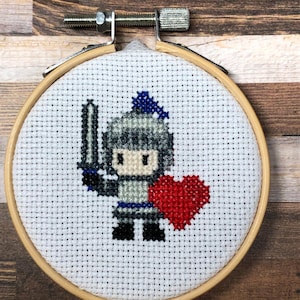 May include: Cross-stitch embroidery of a pixelated knight holding a sword and a red heart. The knight wears gray armor with blue accents. The embroidery is set in a wooden hoop with a metal adjustment screw.