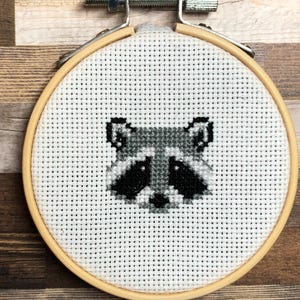 Könnte beinhalten: Kreuzstich-Stickerei eines Waschbärgesichts mit schwarzen und grauen Details auf weißem Hintergrund. Das Design befindet sich in einem runden Stickrahmen aus Holz, der mit einem Metallverschluss befestigt ist.