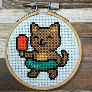 May include: A cross-stitch embroidery in a round wooden hoop featuring a brown cat with a light brown belly, wearing a teal inner tube, and holding an orange ice lolly. The design is stitched on white Aida fabric with a visible grid pattern.