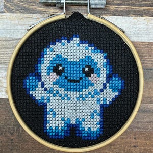 Könnte beinhalten: Eine Kreuzstichstickerei mit einem pixeligen Yeti-Charakter mit weißem und blauem Fell, schwarzen Augen und rosa Wangen, zentriert auf schwarzem Stoff. Das Design befindet sich in einem runden Holzstickrahmen mit einer metallenen Spannschraube.