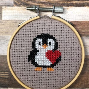Könnte beinhalten: Kreuzstich-Stickerei eines Pinguins, der ein rotes Herz hält, in einem hölzernen Stickrahmen. Der Pinguin ist schwarz, weiß und orange, mit grauem Hintergrund. Der Rahmen wird mit einer Metallschraube befestigt.