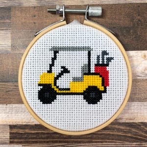 Puede incluir: Un bastidor de bordado de punto de cruz con un carrito de golf amarillo con ruedas negras, un techo gris y una bolsa roja. El carrito de golf está sobre un fondo blanco, enmarcado por un aro de madera. El fondo es una superficie de madera.