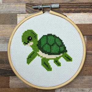 以下が含まれることがあります： 白いアイーダ布にクロスステッチで刺繍された、濃い緑色の甲羅を持つ小さな緑色のカメ。布は、上部に金属製のテンションネジが付いた円形の木製刺繍枠にしっかりと固定されています。