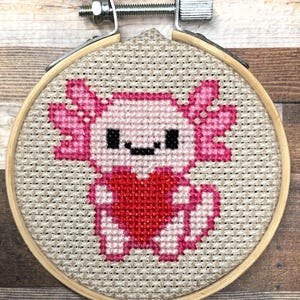Könnte beinhalten: Kreuzstich-Stickerei eines rosa Axolotls, das ein rotes Herz hält. Das Axolotl hat schwarze Augen und einen lächelnden Mund. Die Stickerei befindet sich in einem Holzrahmen mit einem Metallspannmechanismus.