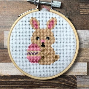 Puede incluir: Bordado de punto de cruz de un conejito sosteniendo un huevo de Pascua. El conejito es marrón con detalles rosas, y el huevo es rosa y morado. El bordado está en un bastidor de madera.