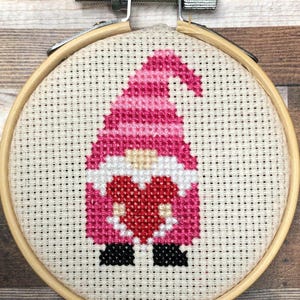 May include: A cross-stitch embroidery of a gnome holding a red heart, set in a wooden embroidery hoop. The gnome's hat and outfit are shades of pink, with a white beard and black shoes. The background is a light beige fabric.