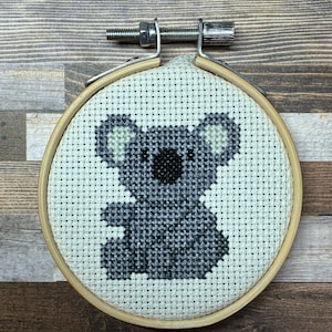 Könnte beinhalten: Eine Kreuzstichstickerei eines grauen Koalas, zentriert in einem runden Stickrahmen aus Holz. Der Koala ist mit grauen und schwarzen Fäden auf cremefarbenem Aida-Stoff gestickt und zeigt eine runde Nase sowie ausgeprägte Ohren.
