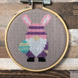 以下が含まれることがあります： 木製のフレームに入ったクロスステッチ刺繍。ウサギの耳を持つノームがイースターエッグを持っています。ノームはピンク、紫、白の衣装に黒い靴を履いています。背景は明るい灰色の生地です。