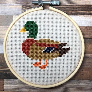 May include: A cross-stitch embroidery of a duck in a wooden hoop. The duck features green, brown, orange, and blue details. The hoop is secured with a metal screw and is displayed on a wooden surface.