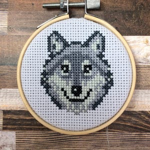 May include: A cross-stitch embroidery of a wolf's head in a wooden hoop. The wolf is rendered in shades of gray and black, with a smiling expression. The hoop is secured with a metal screw and nut.
