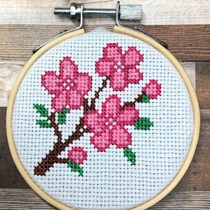 Könnte beinhalten: Drei Keramikbecher mit cremefarbener, gesprenkelter Glasur. Jeder Becher zeigt ein handgemaltes Blumenmuster in Blau-, Grün- und Gelbtönen. Die Becher haben eine schlichte, runde Form und einen bequemen Henkel.