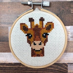 Könnte beinhalten: Eine Kreuzstich-Stickerei eines Giraffenkopfes mit braunen und beigen Fäden auf weißem Stoff. Die Stickerei befindet sich in einem runden Holzreifen mit einem Metallspannmechanismus. Die Giraffe hat schwarze Augen und einen lächelnden Mund.