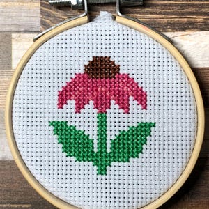 Könnte beinhalten: Kreuzstich-Stickerei einer rosa Sonnenhutblume mit braunem Zentrum und grünem Stiel und Blättern. Die Stickerei befindet sich in einem runden Holzreifen mit einem Metallspannmechanismus. Der Hintergrund ist eine Holzoberfläche.