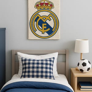 May include: A wall hanging featuring the Real Madrid crest, with a gold crown and a blue and gold emblem. The tapestry hangs above a bed with a blue and white checkered pillow and a soccer ball lamp.
