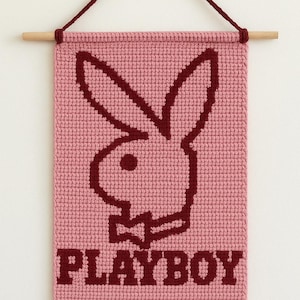 May include: A pink, rectangular wall hanging featuring a burgundy bunny logo and the word "PLAYBOY" in burgundy block letters. The hanging is made of a textured material and is suspended from a wooden dowel with burgundy string.