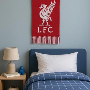 Pode incluir: Uma tapeçaria vermelha com o emblema do Liverpool Football Club, com um pássaro branco e as letras L.F.C. Acima de uma cama com edredão xadrez azul e branco. Uma lâmpada e um robô de brinquedo estão numa mesa de cabeceira de madeira.