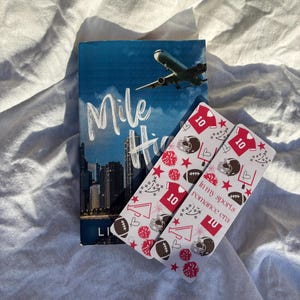 Op de afbeelding: Een boek getiteld "Mile High" met een blauwe omslag met een stadsgezicht en een vliegtuig. Twee boekenleggers met voetbalthema met rode en witte ontwerpen, voetbalhelmen en de tekst "In my sports romance era."