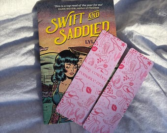May include: A book titled "Swift and Saddled" with a vintage-style cover, accompanied by two pink bookmarks. The bookmarks feature cowboy-themed illustrations and text. The book cover has a woman with blue hair and a cowboy.