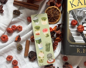 May include: A bookmark featuring watercolor illustrations of various iced drinks, including matcha lattes and frappes, rests on a white cloth. Surrounding the bookmark are decorative elements like a candle, cinnamon sticks, dried orange slices, and small pumpkins.