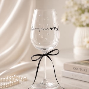 May include: A clear glass wine glass featuring the name Giorgia in black cursive script, accompanied by two small black hearts. The glass is decorated with scattered white dots and tied with a thin black ribbon around the stem.