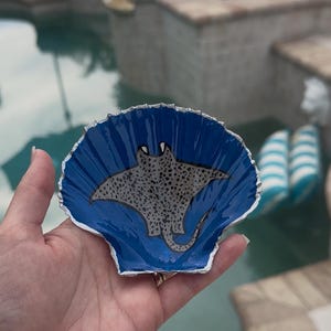 May include: A blue seashell with a silver rim, painted with a black and white manta ray. The seashell is being held in a hand.
