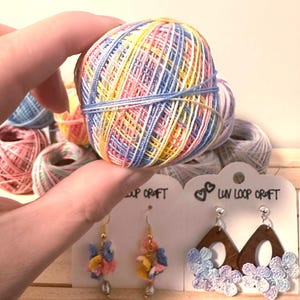 Puede incluir: Una mano sostiene una madeja de hilo multicolor con tonos azules, amarillos, rosas y blancos. En el fondo, hay varias madejas de hilo más. Debajo, dos pares de pendientes con diseños florales y el texto "Luv Loop Craft".