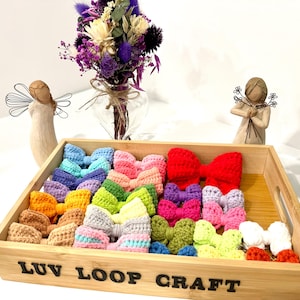 May include: A wooden tray filled with colorful, crocheted hair bows. The bows are in various colors, including red, blue, green, and pink. The tray has the text "Luv Loop Craft" on the front. Decorative angels and a vase of flowers are in the background.