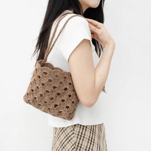 May include: A brown crochet handbag with a scalloped top edge and woven handles. The bag has a geometric openwork design. The bag is carried over the shoulder.