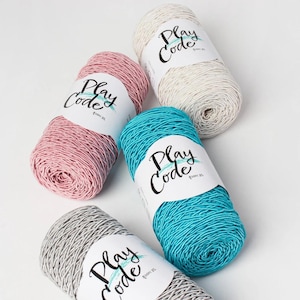May include: Five skeins of yarn in shades of pink, white, teal, grey, and light beige. Each skein features a white label with the words "Play Code" in a handwritten style. The yarn is tightly wound and looks soft.