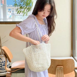 May include: A woman wearing a lilac gingham dress with short sleeves and a ruffled hem, holding a cream-colored, open-weave tote bag with a rope handle. The bag is a light beige color and appears to be made of a woven material.