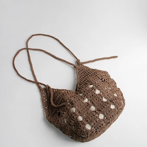 Puede incluir: Un bolso tote de ganchillo marrón de tejido abierto con correas largas y finas. El bolso tiene forma redondeada con una serie de agujeros espaciados uniformemente por todo el cuerpo. Las correas están anudadas en la parte superior del bolso.