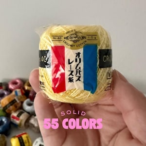 May include: A ball of yellow crochet cotton with a red and blue stripe label. The label has Japanese text and the number 40. The words "SOLID 55 COLORS" are in pink text below the ball of yarn. Other yarn balls are in the background.